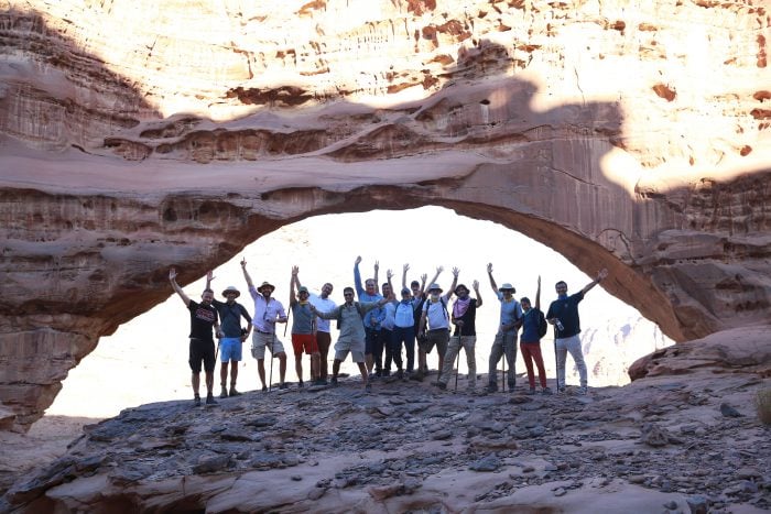 Large group of men and women in summer clothes and their hands in the air stood under a large arched rock