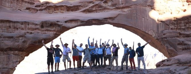 Large group of men and women in summer clothes and their hands in the air stood under a large arched rock