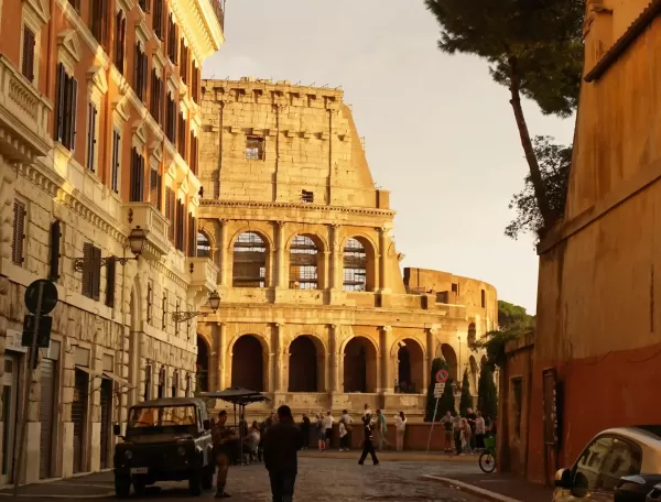 View of the side of the Colosseum from a side street with people buildings and parked car