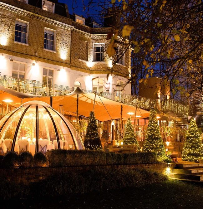 Exterior Riverhouse building lit up with Christmas lights, outside seating dome next to building surrounded by light up Christmas trees.