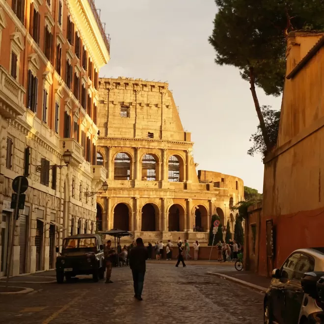View of the side of the Colosseum from a side street with people buildings and parked car